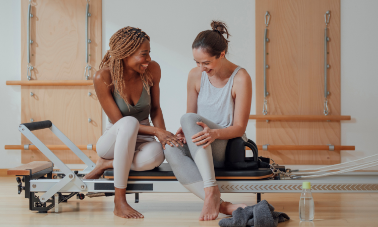 Two pilates students giggle on the reformer after their class led by their teacher who has yoga insurance.