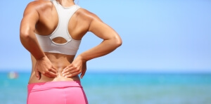 Back pain. Athletic woman in pink sportswear standing at the seaside rubbing the muscles of her lower back, cropped torso portrait.